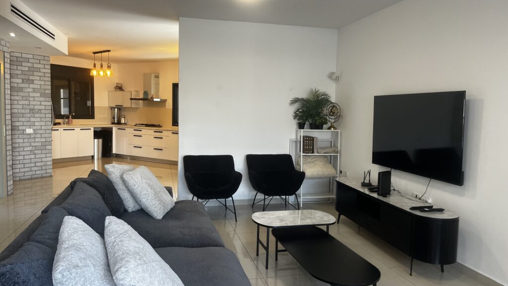Spacious living room with modern gray sofa, flat-screen TV, and open kitchen in a luxury villa in Netanya.
