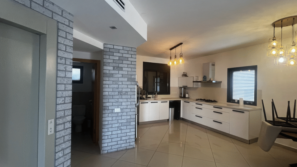 Modern open-plan kitchen in a luxury Netanya apartment featuring white cabinetry, elegant lighting fixtures, and contemporary gray brick walls