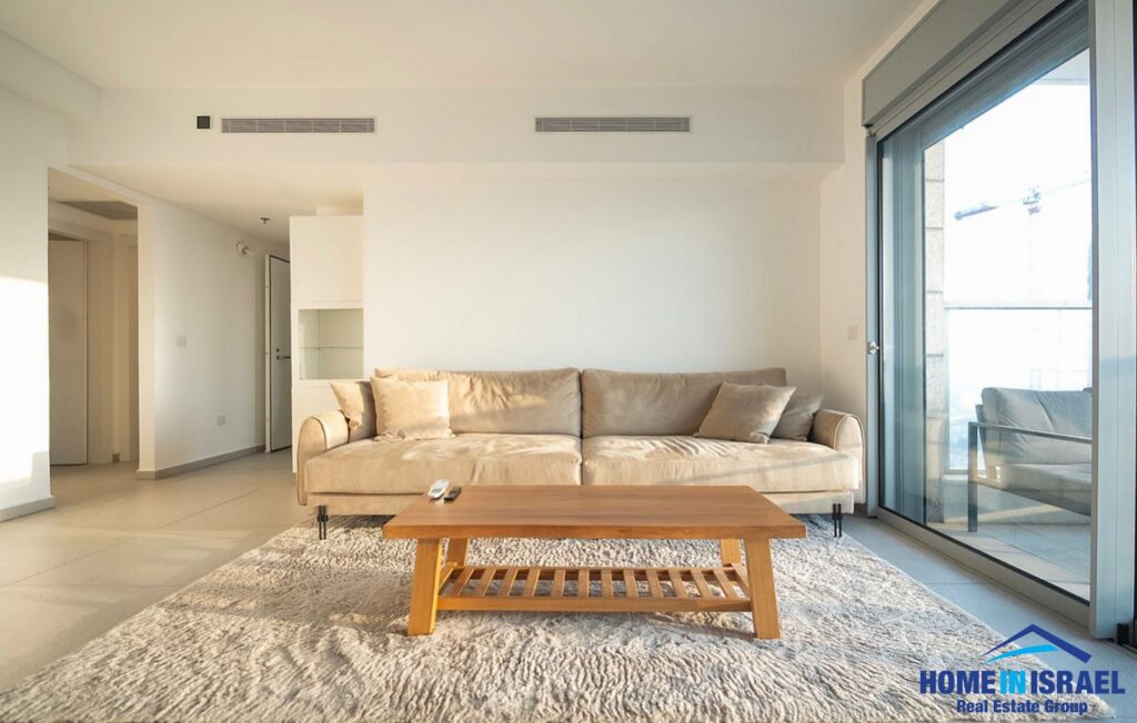 Modern, sunlit living room interior featuring a beige sofa, a wooden coffee table on a shaggy rug, and large glass sliding doors opening to a high-floor balcony in Netanya.